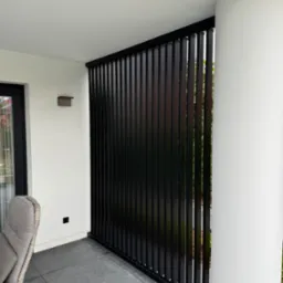 Nowoczesny balkon z czarną, pionową osłoną przeciwsłoneczną i szarymi płytkami podłogowymi, widok z boku na kolumnę i krzesło z rattanu.