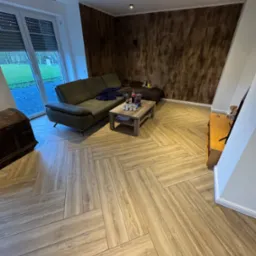 Salon z drewnopodobną podłogą ułożoną we wzór jodełki, zieloną sofą i ścianą z efektem drewna.