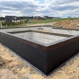 Staszel Bau - Mateusz Staszel - Zabezpieczone fundamenty domu z bloczków betonowych, pomalowane na czarno, z widoczną izolacją. W tle domy w budowie i koparka.