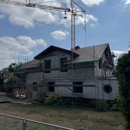 Staszel Bau - Mateusz Staszel - Budowa domu z cegły silikatowej w trakcie realizacji. Widoczne rusztowania, częściowo położony dach z czerwonej dachówki i żuraw budowlany w tle.
