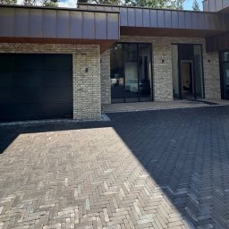 Staszel Bau - Mateusz Staszel - Nowoczesny dom z elewacją z jasnej cegły, ciemną bramą garażową i brukowanym podjazdem w jodełkę. Widoczne duże okna i metalowe elementy wykończeniowe.