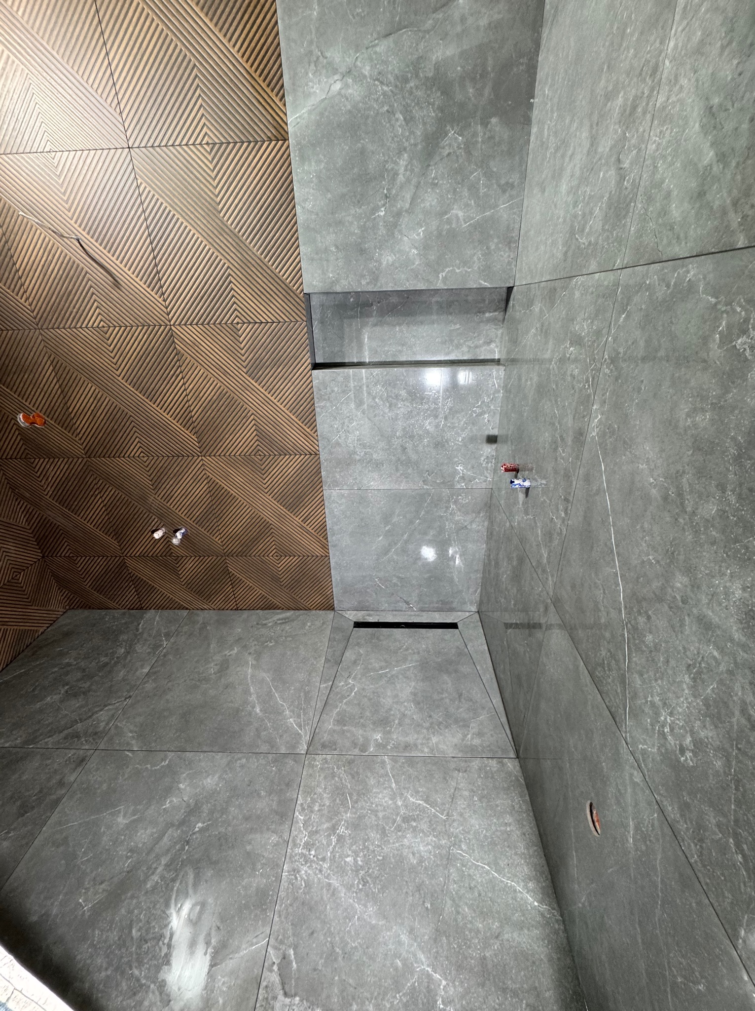 Wnętrze łazienki w trakcie wykańczania, widok z dołu na sufit i ściany wyłożone szarymi płytkami imitującymi marmur, z fragmentem sufitu wykończonym drewnopodobną płytką z geometrycznym wzorem...