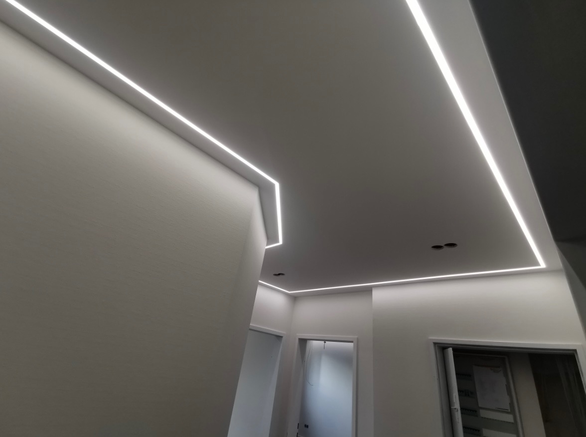 Nowoczesne oświetlenie LED wpuszczone w sufit, tworzące geometryczne linie światła w jasnym wnętrzu z widocznymi drzwiami.