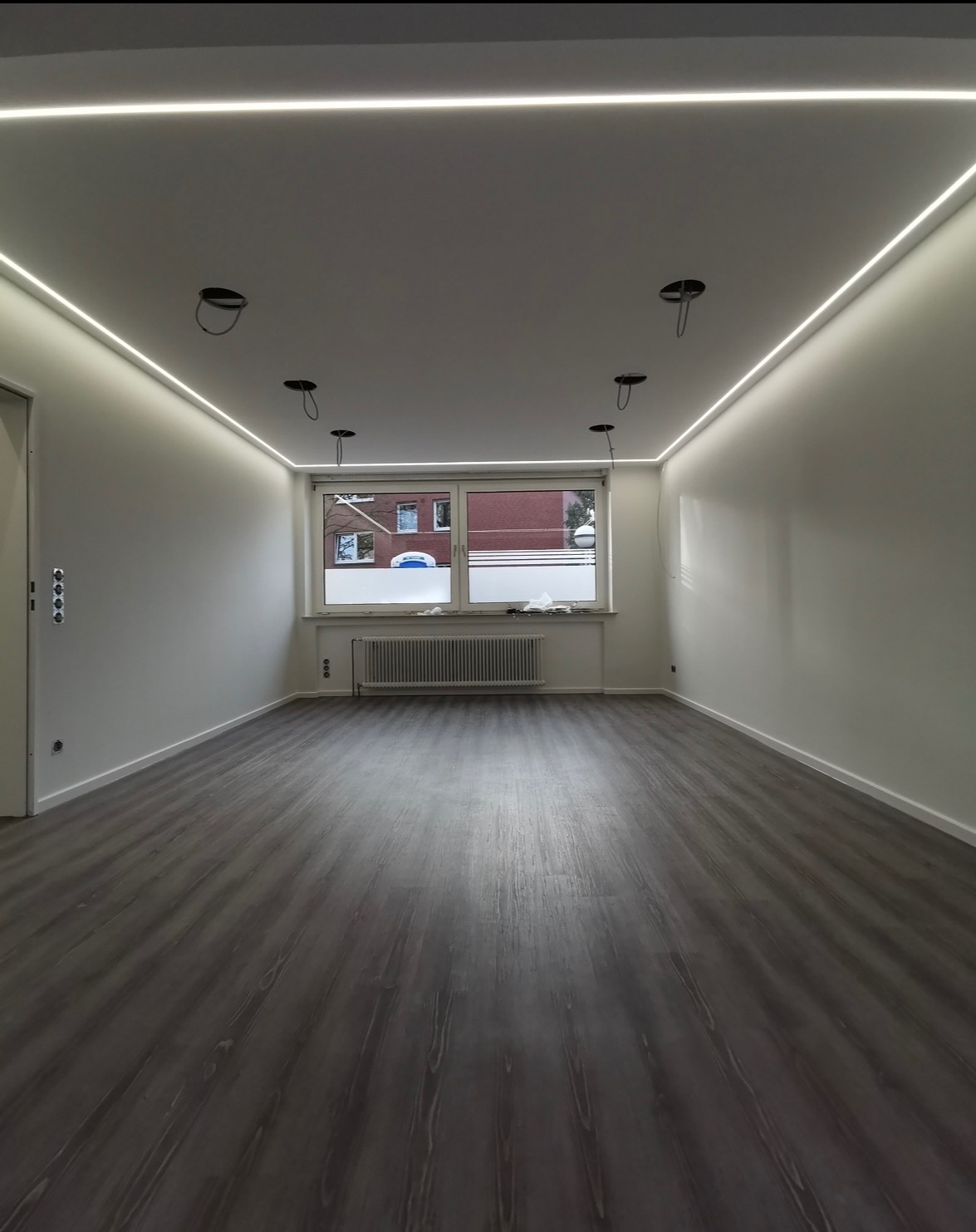 Pusty pokój po remoncie z panelami podłogowymi w odcieniach szarości, białymi ścianami, oświetleniem LED podsufitowym i przygotowanymi otworami na lampy sufitowe.