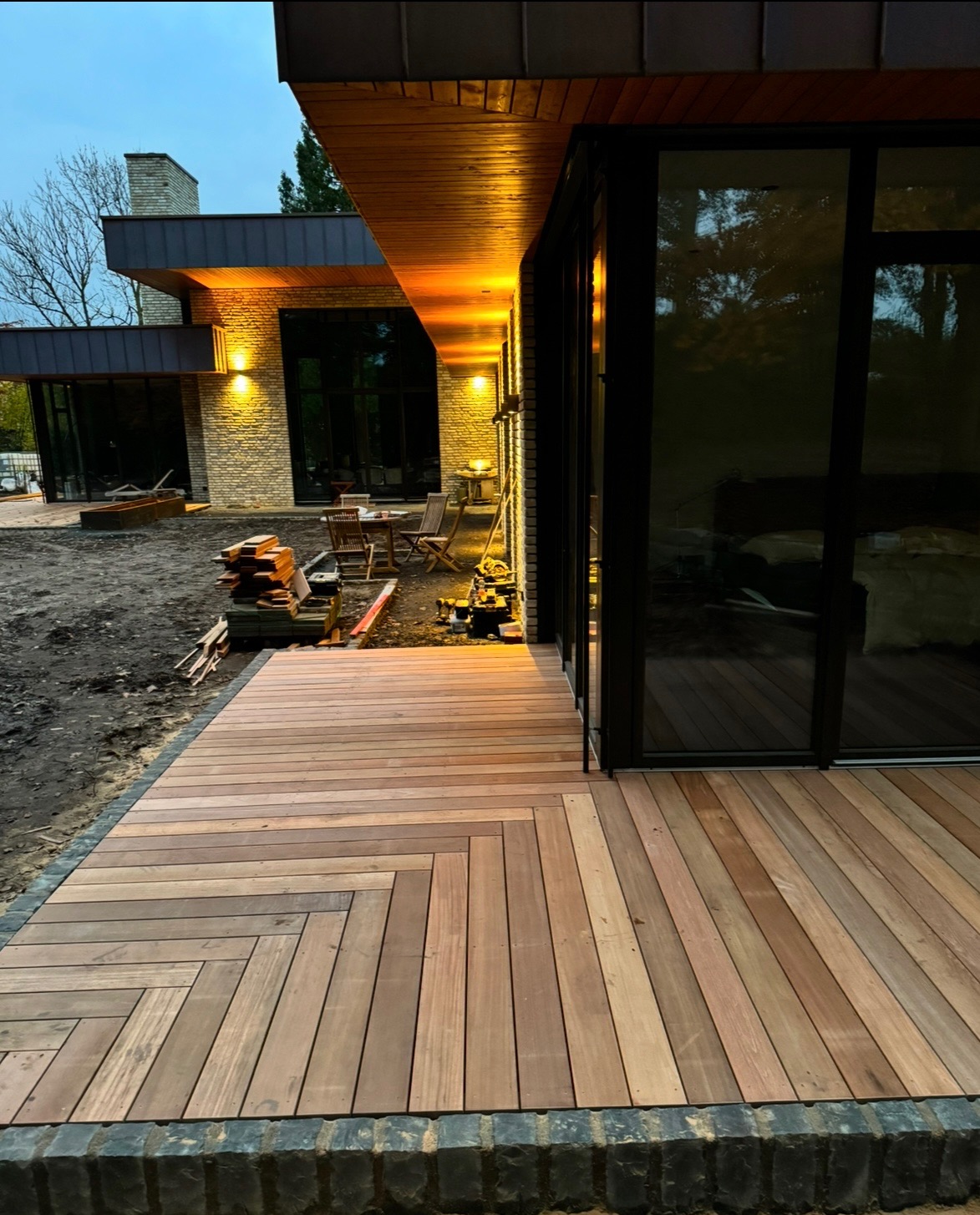 Nowoczesny dom z drewnianym tarasem ułożonym w jodełkę, widoczne narzędzia i materiały budowlane, oświetlenie elewacyjne, wieczorny krajobraz.