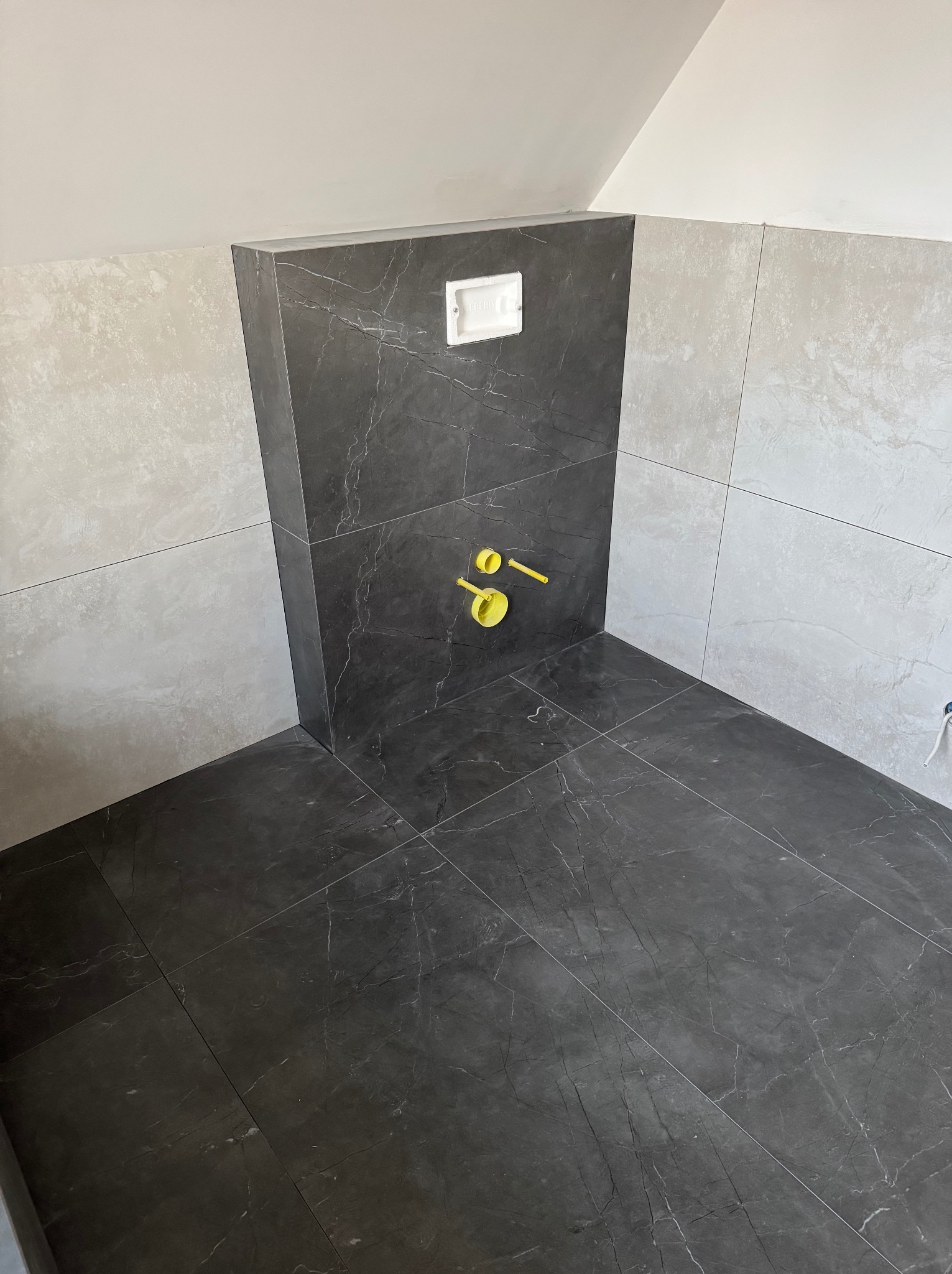 Wykończona łazienka z szarymi płytkami imitującymi marmur na ścianach i podłodze, widoczne żółte rury instalacji wodnej i biały przycisk spłuczki.