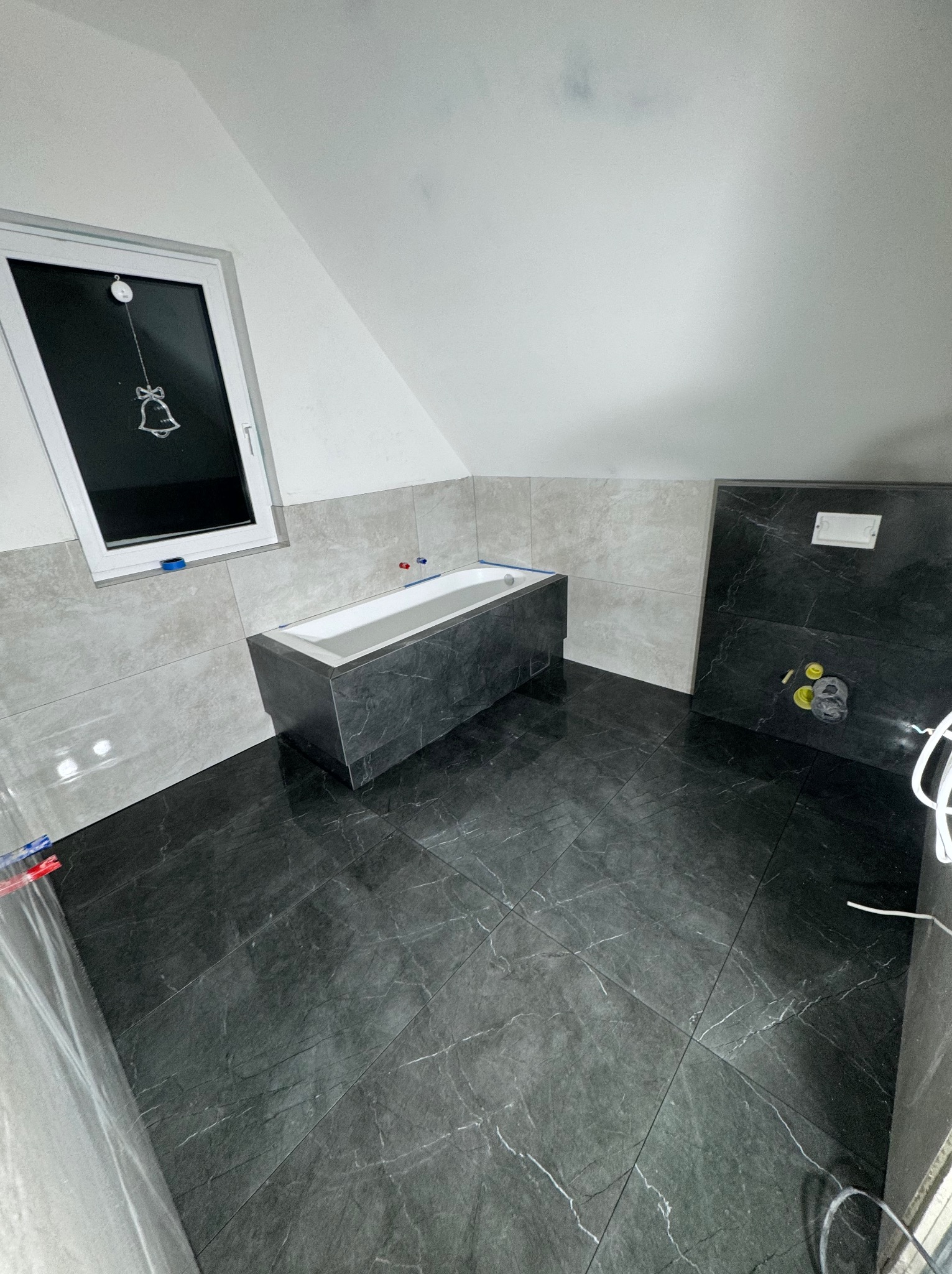 Nowoczesna łazienka w trakcie wykończenia, z czarną marmurową podłogą i obudową wanny, jasnoszare płytki ścienne do połowy wysokości, okno dachowe z dekoracją dzwonka.