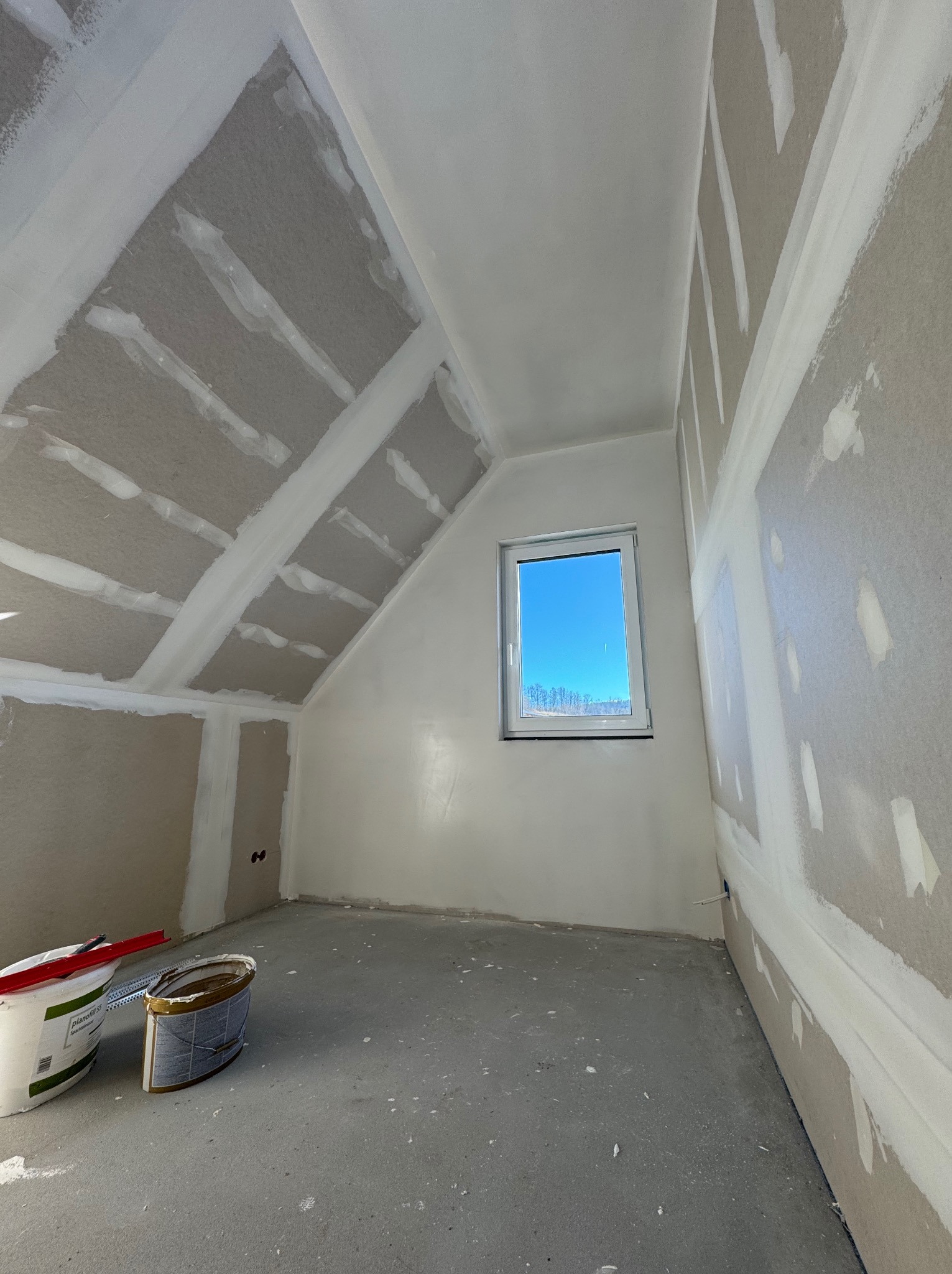Pokój na poddaszu w trakcie prac wykończeniowych: ściany z płyt gipsowo-kartonowych ze szpachlowanymi łączeniami, widoczne dwa wiadra z masą szpachlową i szpachelka, okno z widokiem na błękitne niebo.