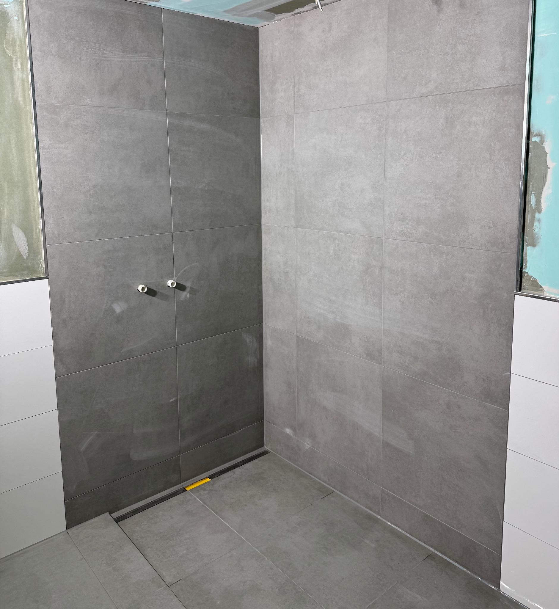 Szare, matowe płytki ceramiczne ułożone w kabinie prysznicowej z odpływem liniowym, widoczne białe rurki wystające ze ściany oraz fragmenty niezakończonych prac wykończeniowych na ścianach.