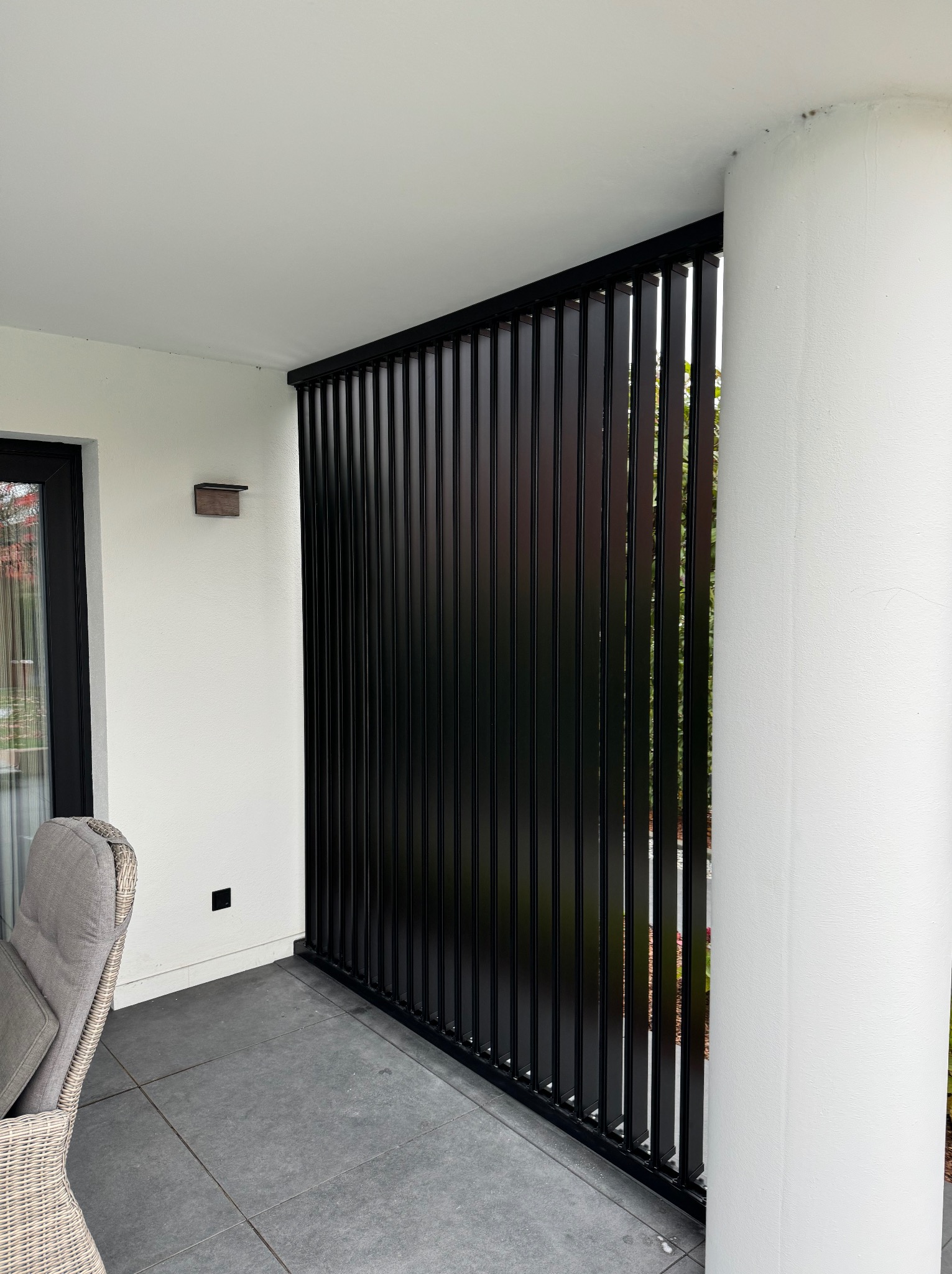 Nowoczesny balkon z czarną, pionową osłoną przeciwsłoneczną i szarymi płytkami podłogowymi, widok z boku na kolumnę i krzesło z rattanu.