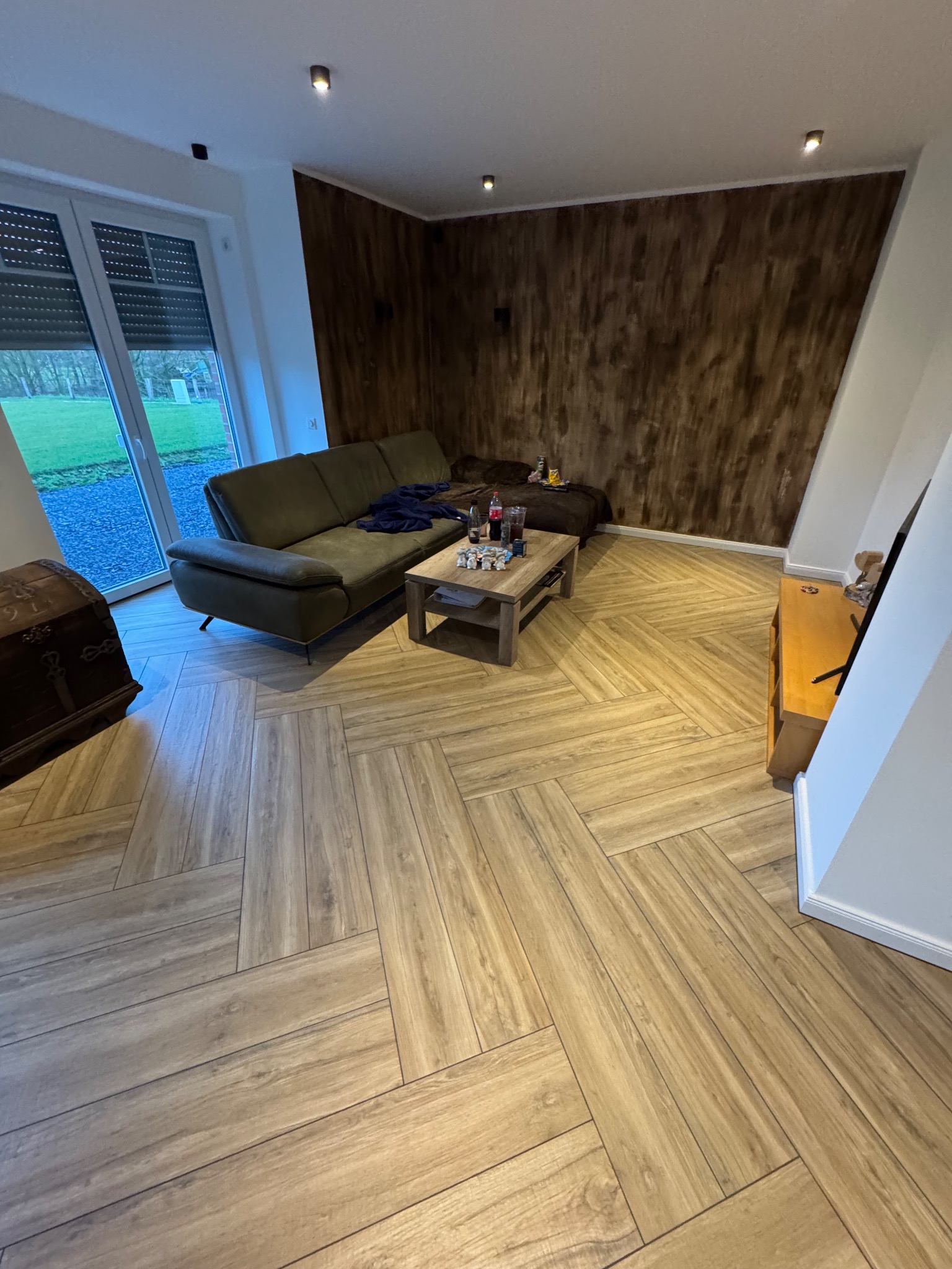 Salon z drewnopodobną podłogą ułożoną we wzór jodełki, zieloną sofą i ścianą z efektem drewna.