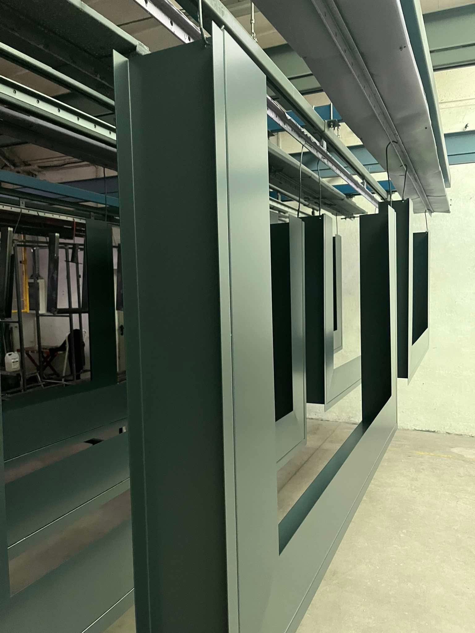 Metalowe ramy okienne zawieszone na wieszakach po procesie malowania, gotowe do wyschnięcia, widoczne ślady po zawieszeniu.