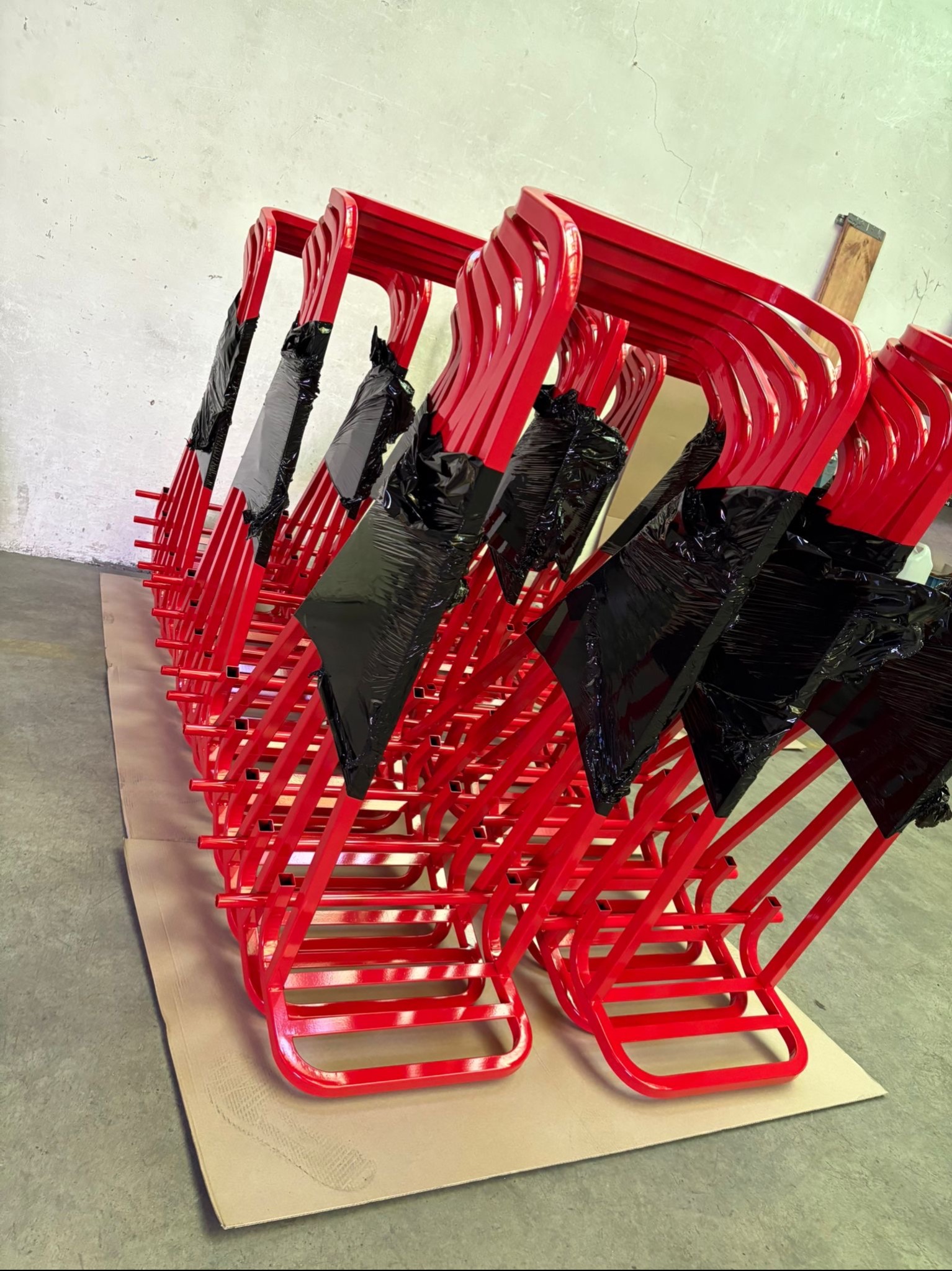 Rząd czerwonych, metalowych stojaków na choinkę, częściowo owiniętych czarną folią, po malowaniu proszkowym, ułożonych na kartonie.