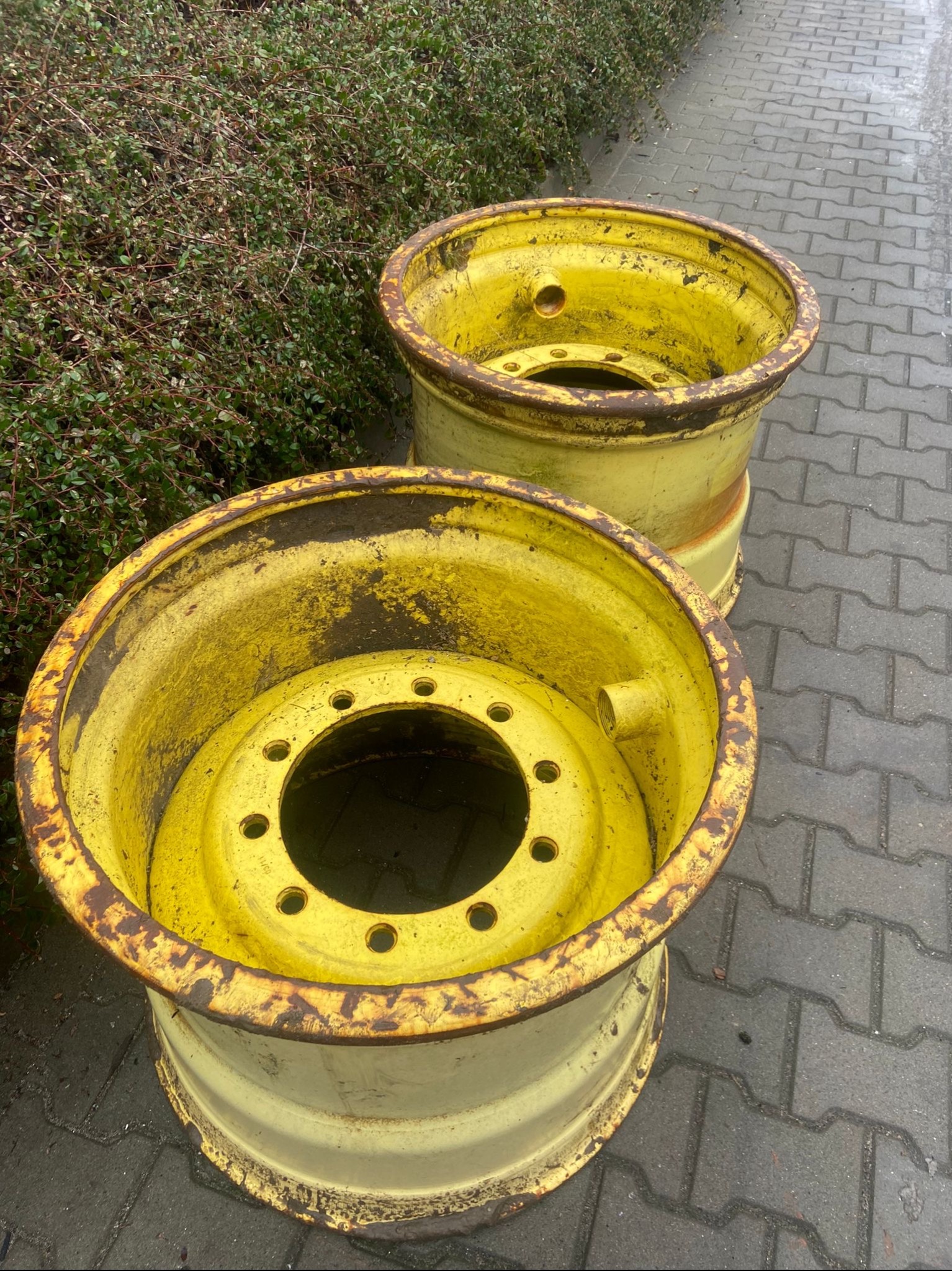 Dwa żółte, metalowe koła do traktora z widocznymi śladami rdzy i zużycia, stojące na szarej kostce brukowej obok krzewu.