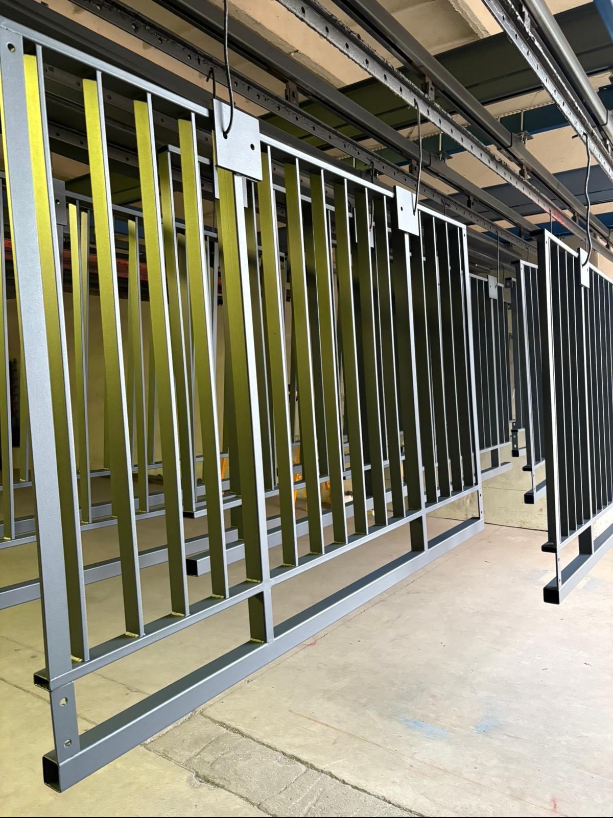 Części metalowe ogrodzenia po malowaniu proszkowym, zawieszone na linii produkcyjnej, niektóre w kolorze żółto-zielonym, inne czarne, w tle betonowa podłoga i sufit hali.