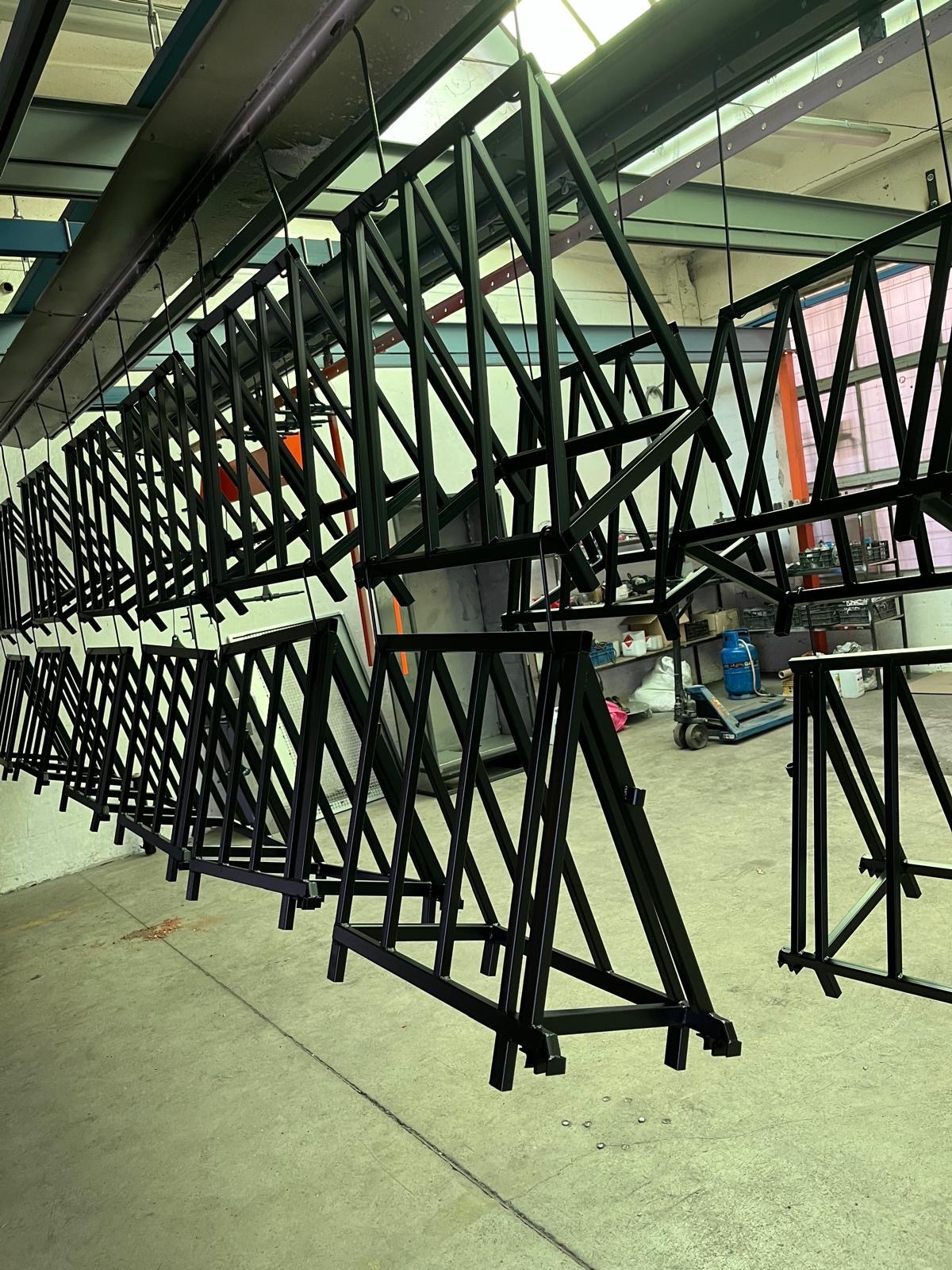 Metalowe konstrukcje w kształcie litery A, zawieszone na hakach po procesie lakierowania, widoczne w hali produkcyjnej.
