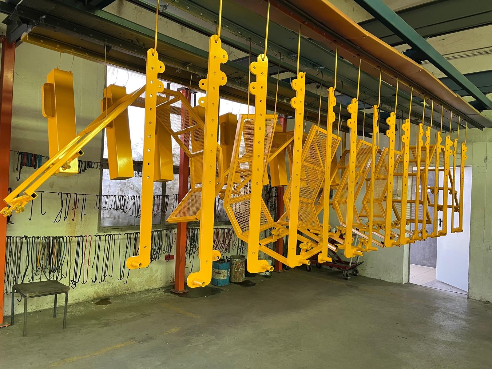 Żółte, metalowe elementy konstrukcyjne, prawdopodobnie kraty lub ramy, zawieszone na hakach po procesie malowania, widoczna surowa hala produkcyjna z oknem i narzędziami w tle.