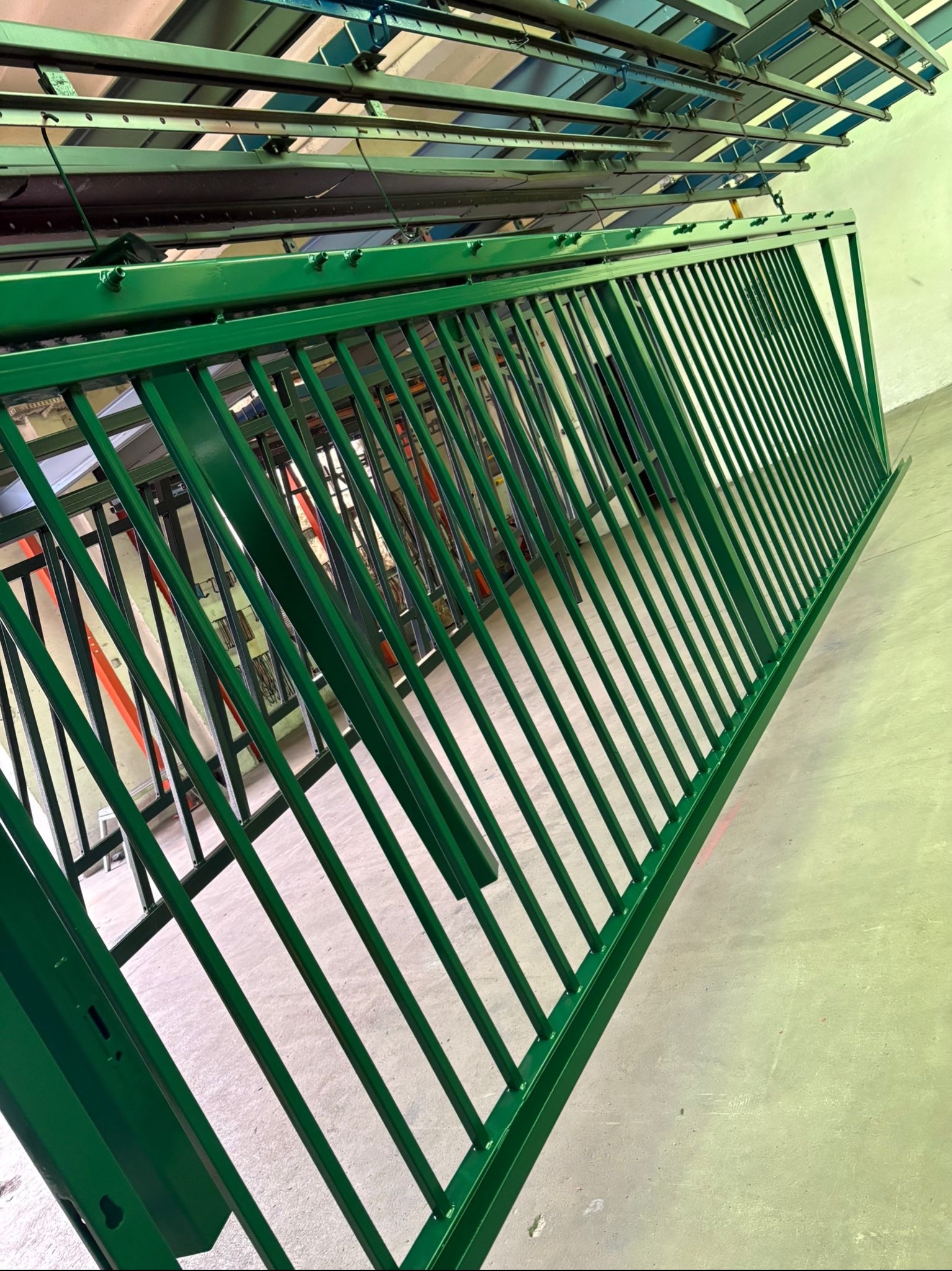 Świeżo pomalowane na zielono, metalowe przęsło ogrodzeniowe zawieszone na systemie transportowym w hali produkcyjnej, gotowe do dalszej obróbki lub transportu.