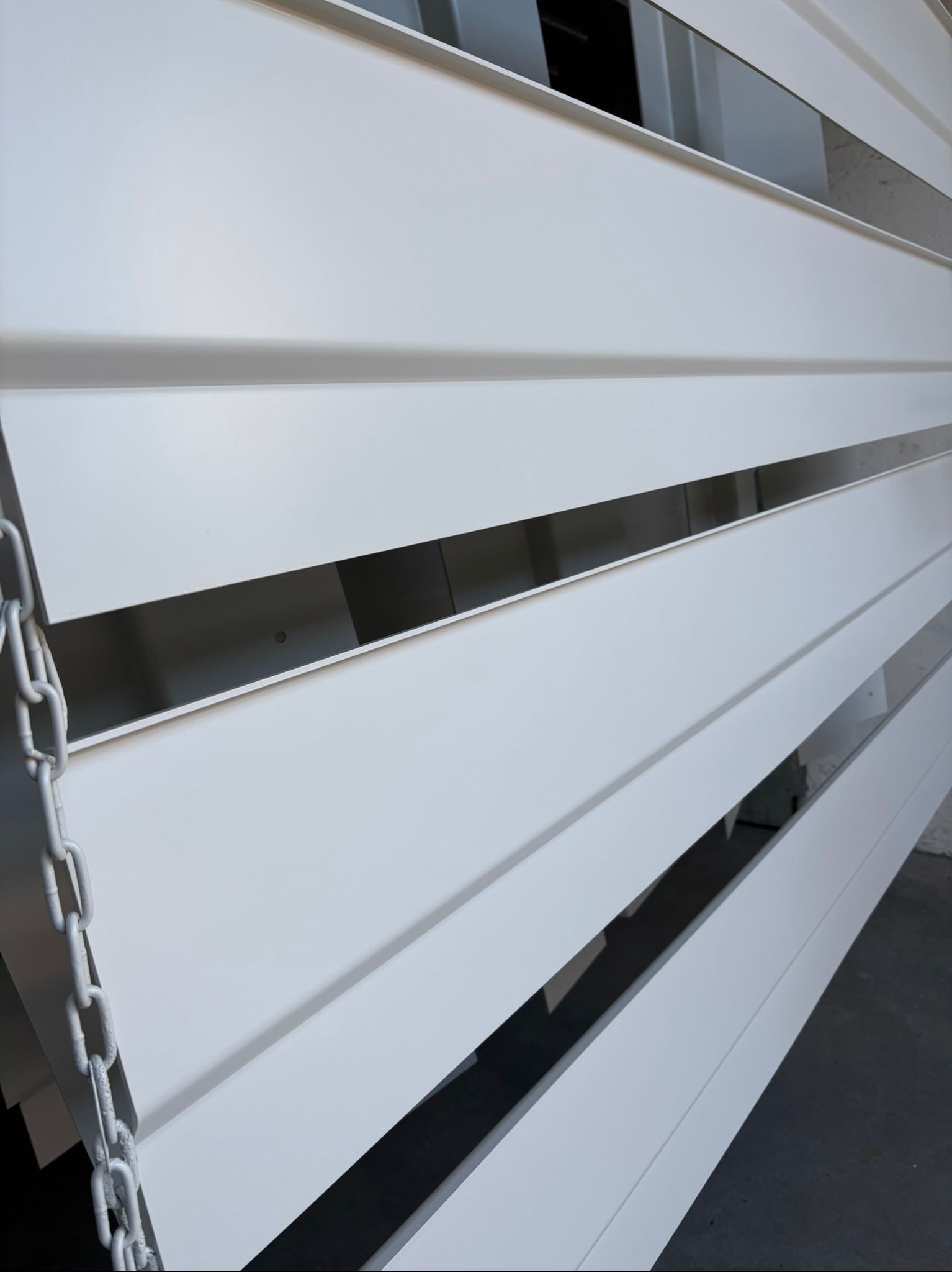 Białe, metalowe żaluzje elewacyjne z łańcuchem po lewej stronie, widoczne z bliska pod kątem.