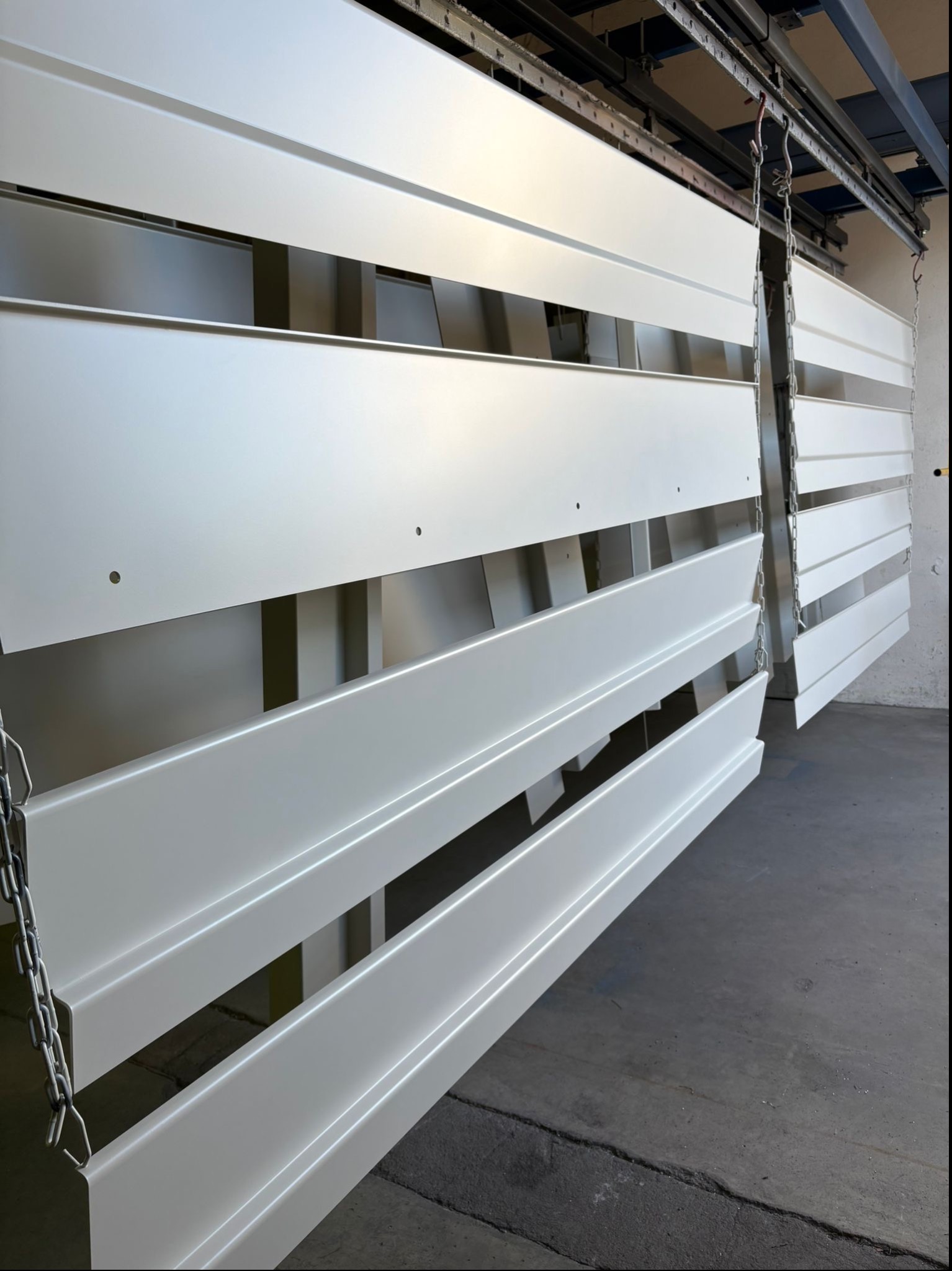 Białe, metalowe elementy zawieszone na łańcuchach podczas procesu malowania, widoczne otwory montażowe, oświetlenie przemysłowe.