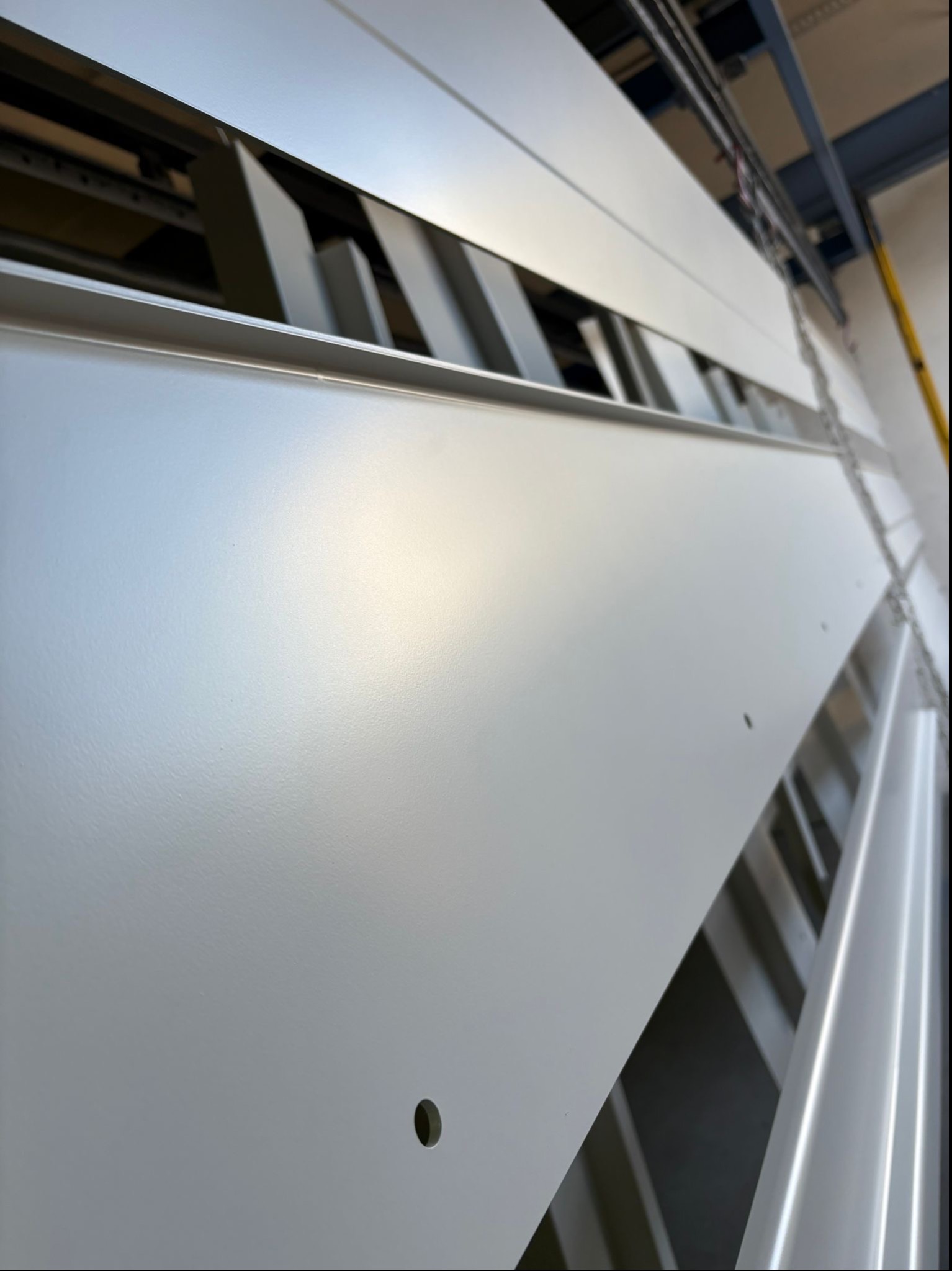 Elementy metalowe zawieszone na łańcuchach, świeżo po malowaniu proszkowym, w odcieniach szarości i bieli, z widocznymi otworami montażowymi.