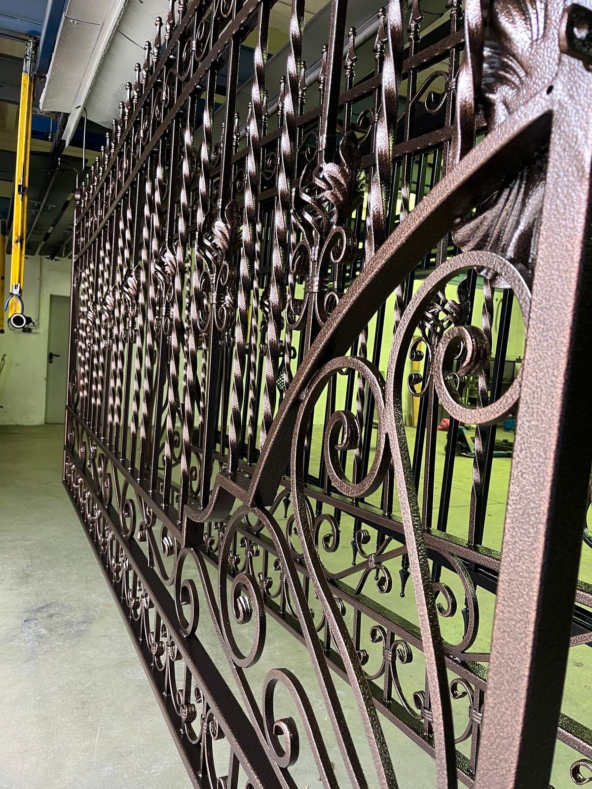 Długa, ozdobna, metalowa brama z kutymi elementami, świeżo po malowaniu proszkowym na brązowo, zawieszona w hali produkcyjnej.
