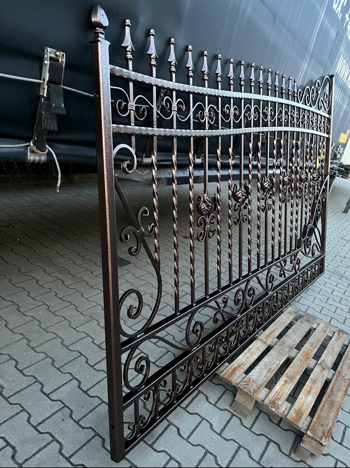 Brama metalowa z ozdobnymi elementami, świeżo po malowaniu, oparta o paletę na tle ciemnej naczepy ciężarówki.