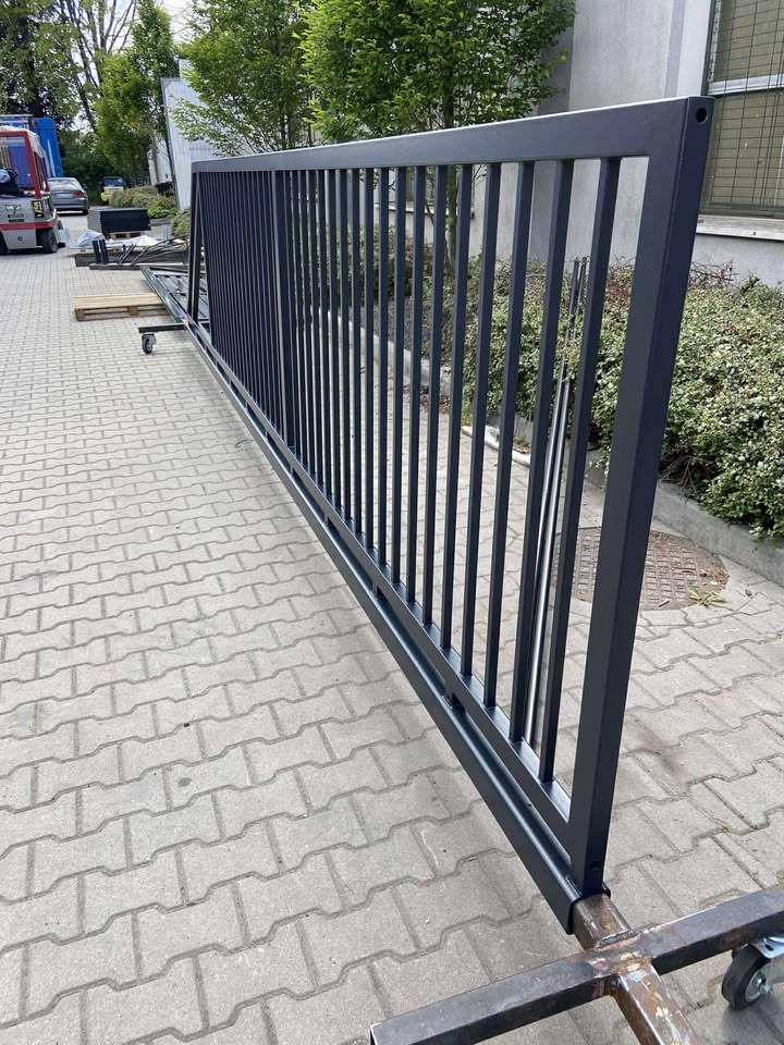 Metalowa, przesuwna brama w kolorze grafitowym, na kółkach, z pionowymi prętami, na tle kostki brukowej i zieleni.