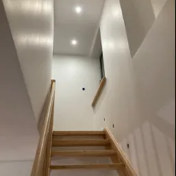 Klatka schodowa z drewnianymi schodami i świeżo pomalowanymi na biało ścianami, widoczne otwory na punkty oświetleniowe i gładkie wykończenie powierzchni.