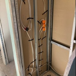 Kamil Górnik BAU - Ściana w trakcie budowy z widocznymi przewodami elektrycznymi przymocowanymi czarnymi opaskami do metalowego stelażu, pomarańczowe puszki instalacyjne widoczne w górnej części, na podłodze zwinięty...