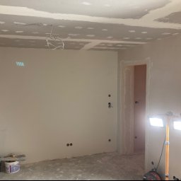 Kamil Górnik BAU - Wnętrze pokoju w trakcie prac wykończeniowych, widoczne ślady szpachlowania na suficie i ścianach, przygotowane otwory pod gniazdka elektryczne, oświetlenie halogenowe na statywie, wiadro z zaprawą...