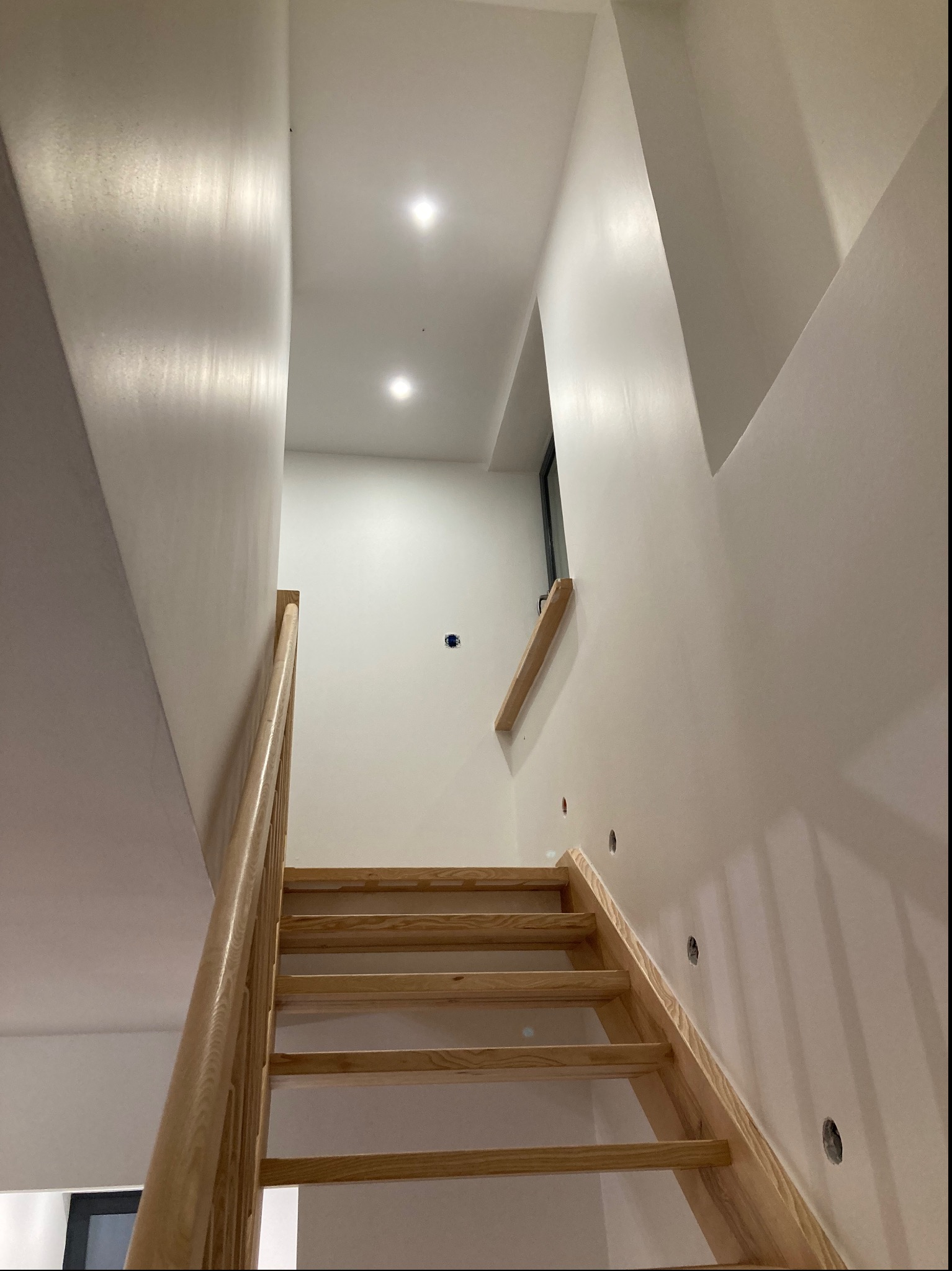 Klatka schodowa z drewnianymi schodami i świeżo pomalowanymi na biało ścianami, widoczne otwory na punkty oświetleniowe i gładkie wykończenie powierzchni.