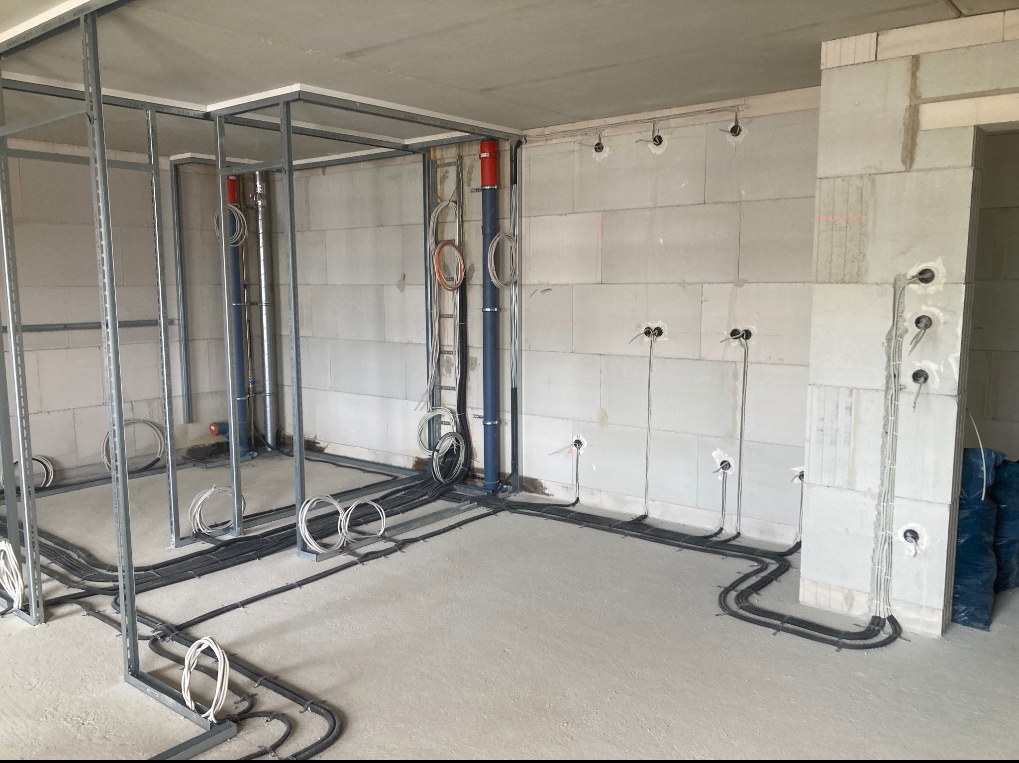 Surowy stan wnętrza z widocznymi instalacjami elektrycznymi w rurach i przewodach, metalowa konstrukcja szkieletowa pod sufitem, ściany z betonu komórkowego z przygotowanymi otworami na gniazdka.