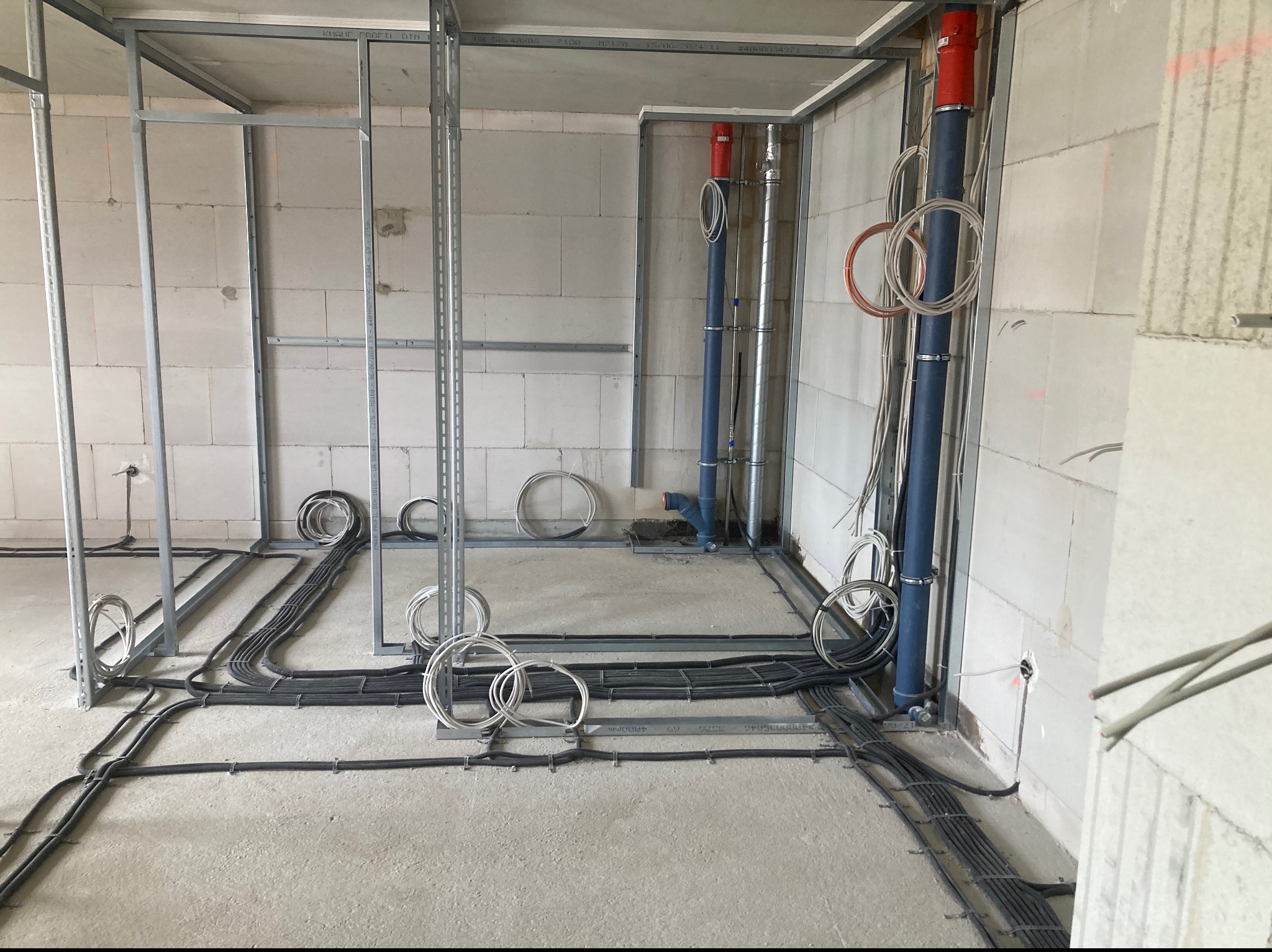 Surowy stan wnętrza z widoczną instalacją elektryczną w peszlach na podłodze, pionowe rury i metalowa konstrukcja ścian działowych.