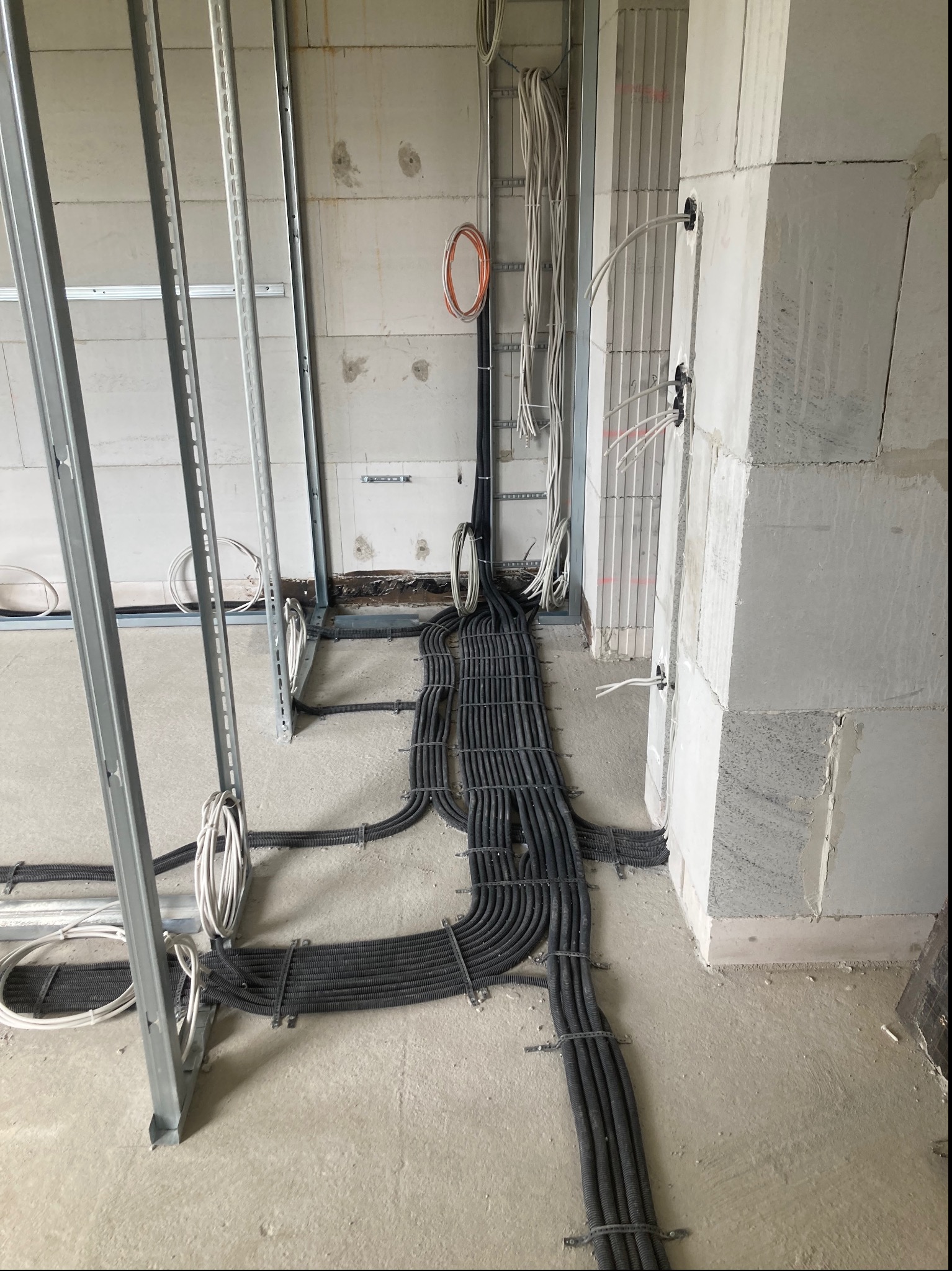 Wiązki czarnych rur karbowanych z kablami, ułożone na podłodze w surowym stanie, prowadzące do ścian z betonu komórkowego z widocznymi puszkami elektrycznymi i wystającymi kablami, oraz metalowe...