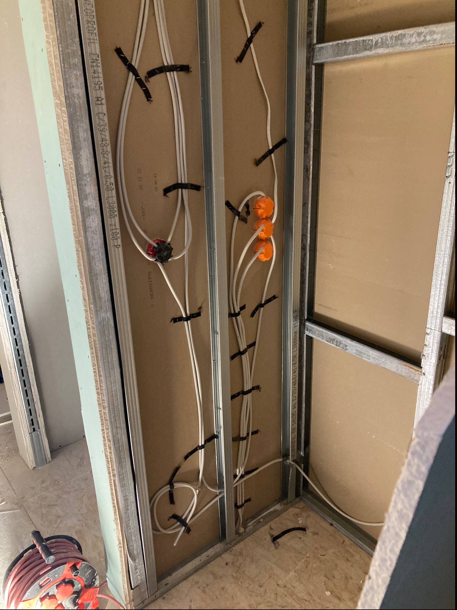 Ściana w trakcie budowy z widocznymi przewodami elektrycznymi przymocowanymi czarnymi opaskami do metalowego stelażu, pomarańczowe puszki instalacyjne widoczne w górnej części, na podłodze zwinięty...