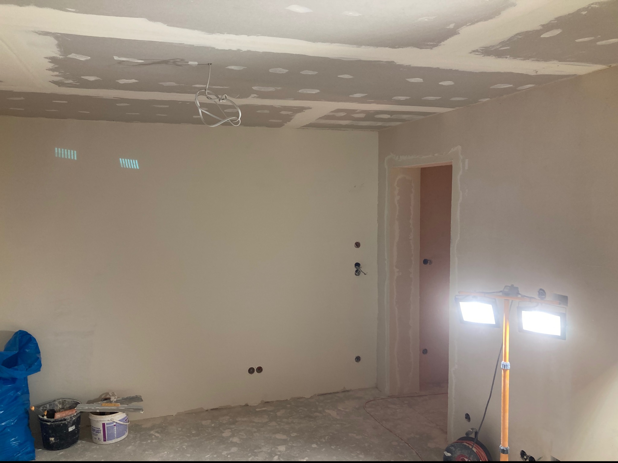 Wnętrze pokoju w trakcie prac wykończeniowych, widoczne ślady szpachlowania na suficie i ścianach, przygotowane otwory pod gniazdka elektryczne, oświetlenie halogenowe na statywie, wiadro z zaprawą...