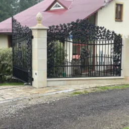 Kuta, czarna brama wjazdowa z motywem roślinnym, osadzona na jasnych, kamiennych słupkach z dekoracyjnymi zwieńczeniami w kształcie szyszki, prowadząca do domu z czerwoną dachówką i kamiennym...