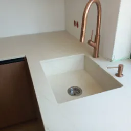 Beżowy blat kamienny z wbudowanym zlewozmywakiem i miedzianą baterią, widok z góry. Minimalistyczny design, subtelny wzór kamienia.