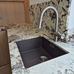 Bella Pietra - Jasny blat kuchenny z naturalnego kamienia o wyrazistym, ciemnym użyleniu, zintegrowany z ciemnym zlewem i nowoczesną baterią, obok drewnianej zabudowy z gniazdkami.