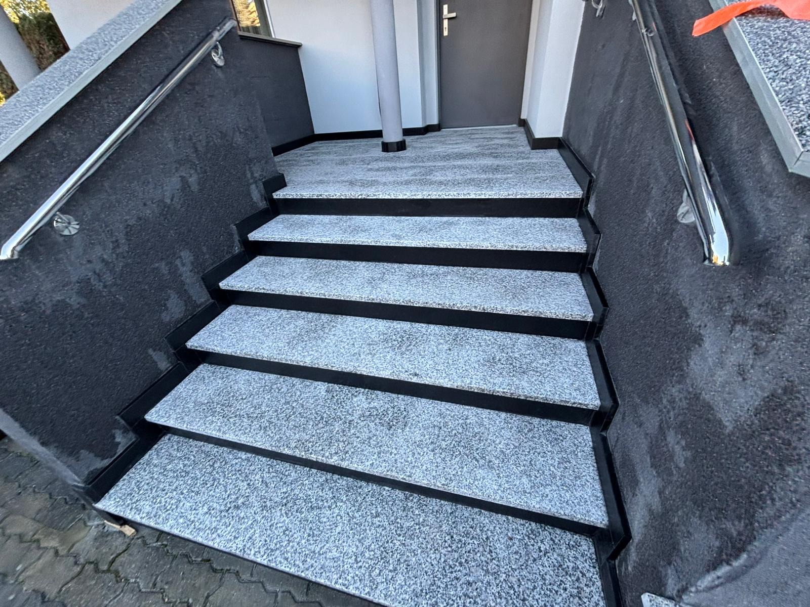 Zewnętrzne schody wejściowe z szarego kamienia z czarnymi obramowaniami stopni i chromowanymi poręczami, prowadzące do drzwi wejściowych.