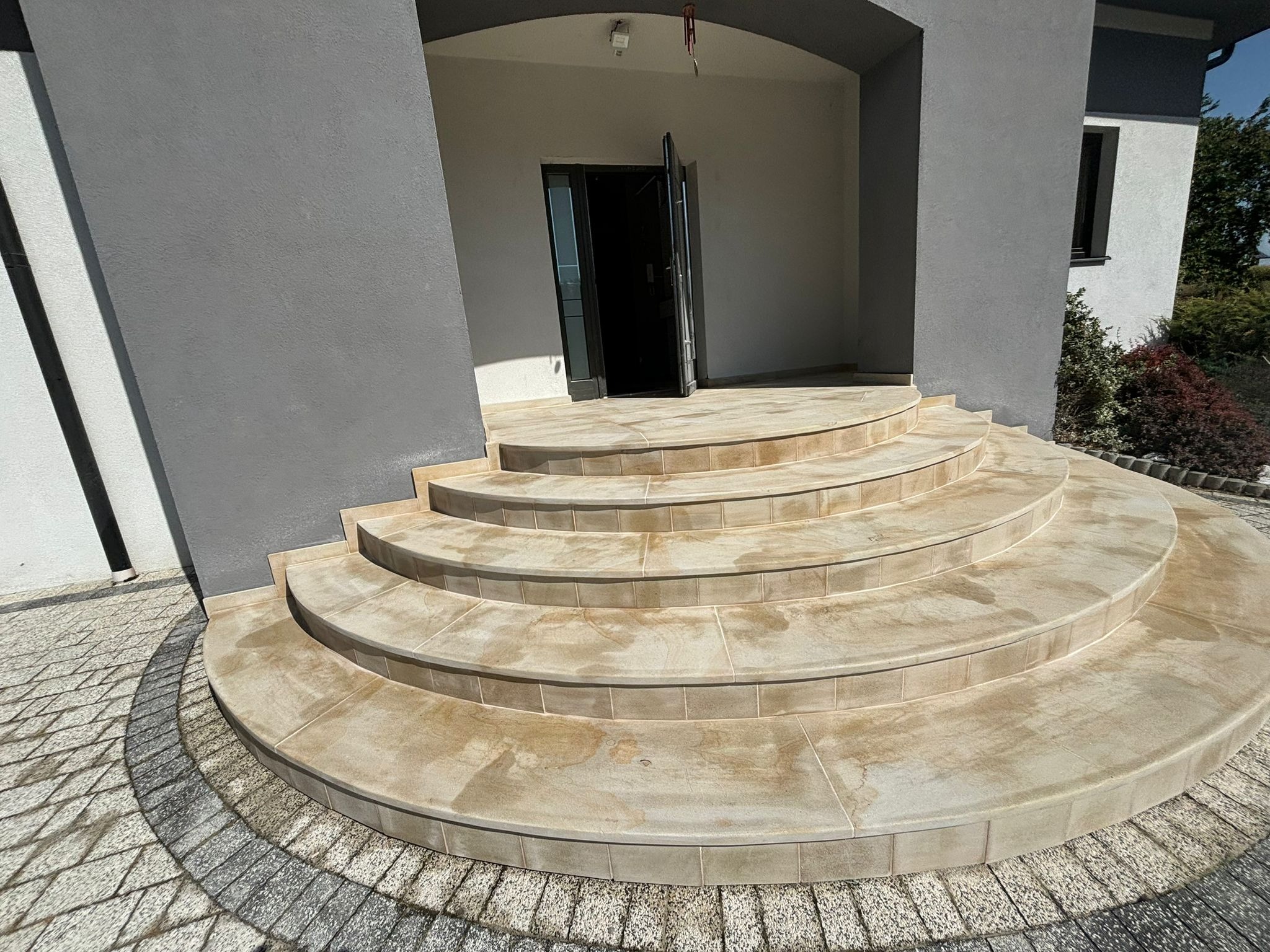 Półokrągłe schody zewnętrzne z jasnych płytek, prowadzące do wejścia domu z otwartymi drzwiami, otoczone szarą kostką brukową.