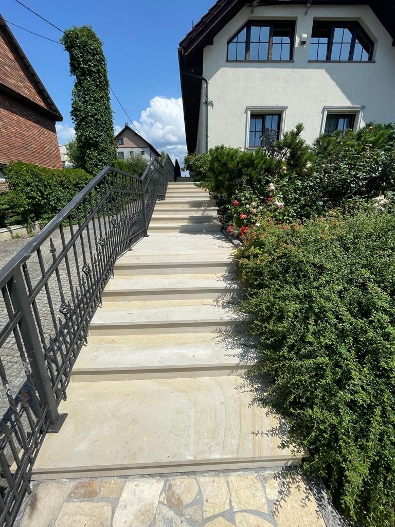 Zewnętrzne schody prowadzące do domu, z metalową balustradą o ozdobnym wzorze, otoczone zielenią i kwitnącymi krzewami róż.