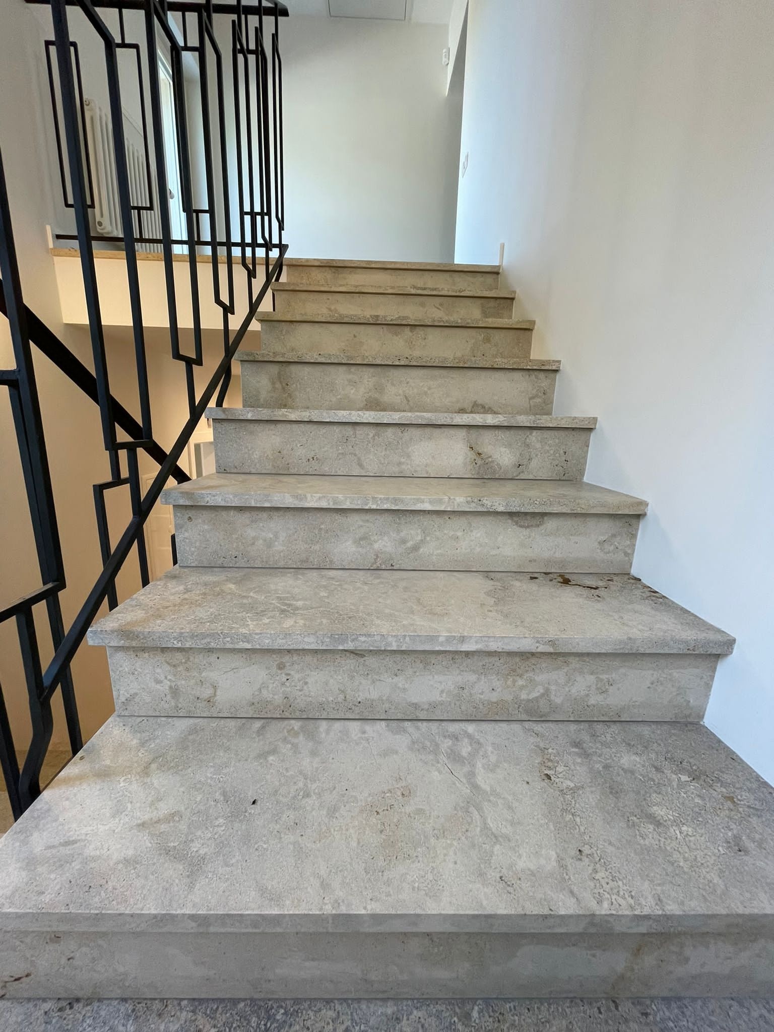 Wewnętrzne schody z jasnego kamienia o porowatej strukturze, z czarną, minimalistyczną balustradą, widok od dołu ku górze.