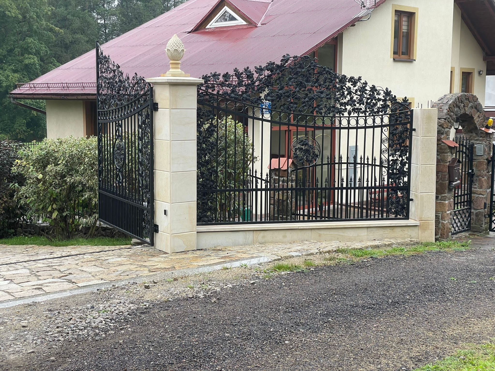 Kuta, czarna brama wjazdowa z motywem roślinnym, osadzona na jasnych, kamiennych słupkach z dekoracyjnymi zwieńczeniami w kształcie szyszki, prowadząca do domu z czerwoną dachówką i kamiennym...