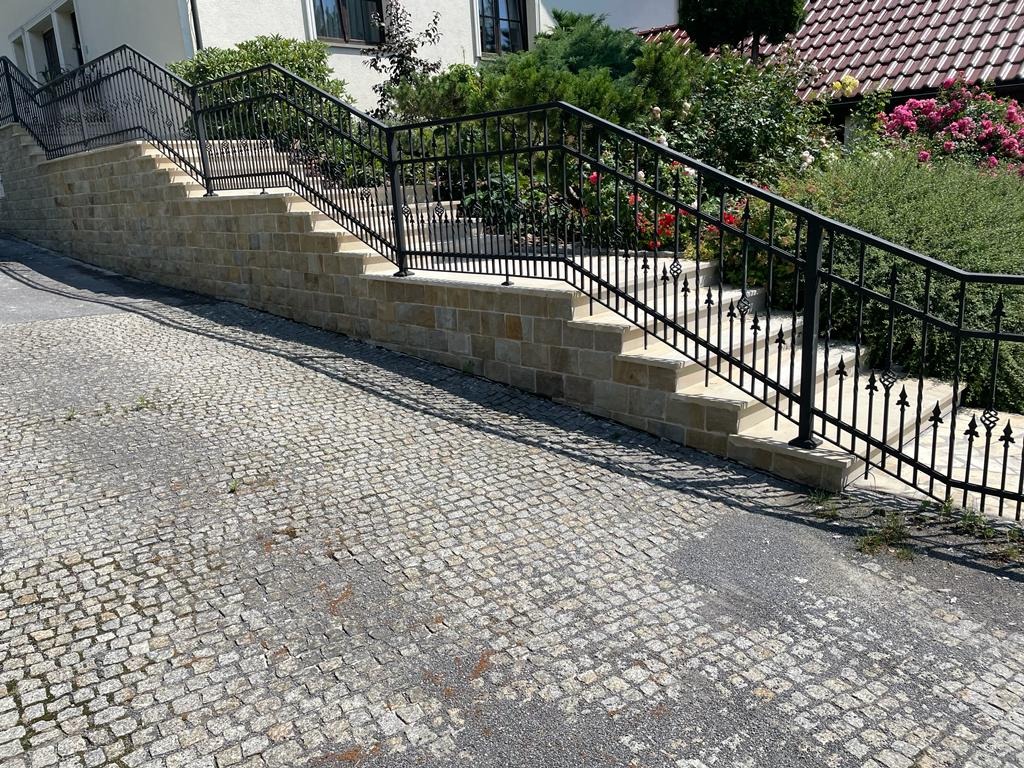 Zewnętrzne schody z jasnego kamienia z czarną, kutą balustradą prowadzące do domu z ogrodem, widok z dołu na nierówną, kamienną nawierzchnię.