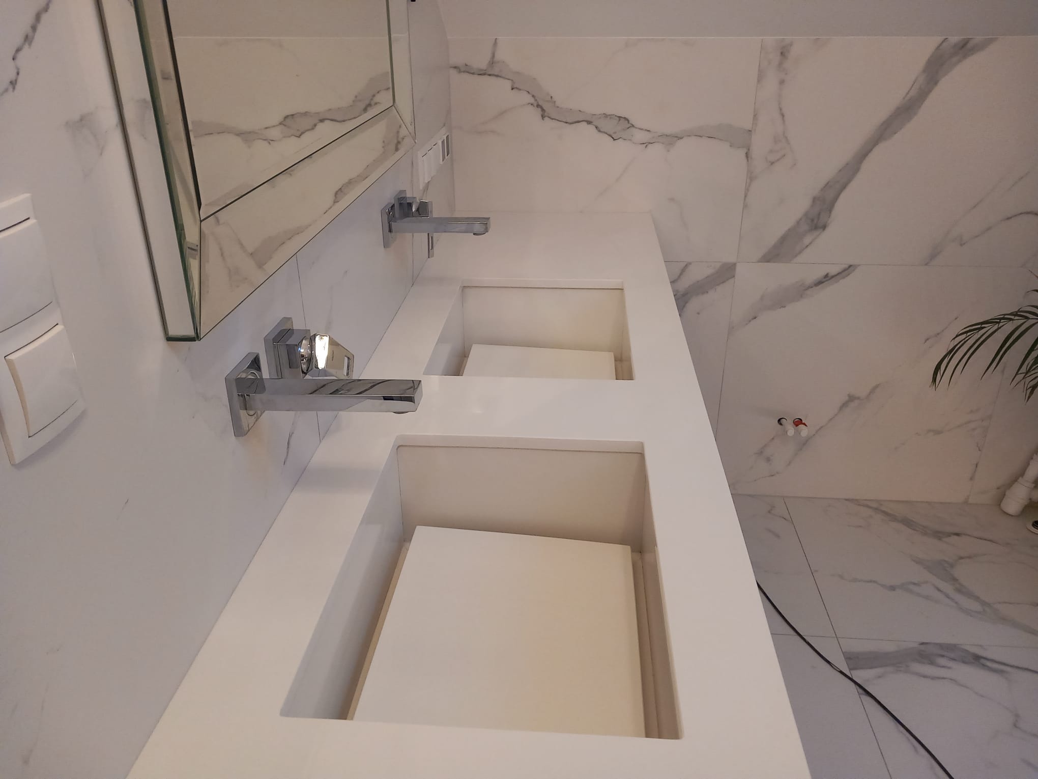 Biała, podwójna umywalka z blatem z konglomeratu na tle marmurowej ściany. Widoczne nowoczesne baterie i fragment lustra. Minimalistyczny design.