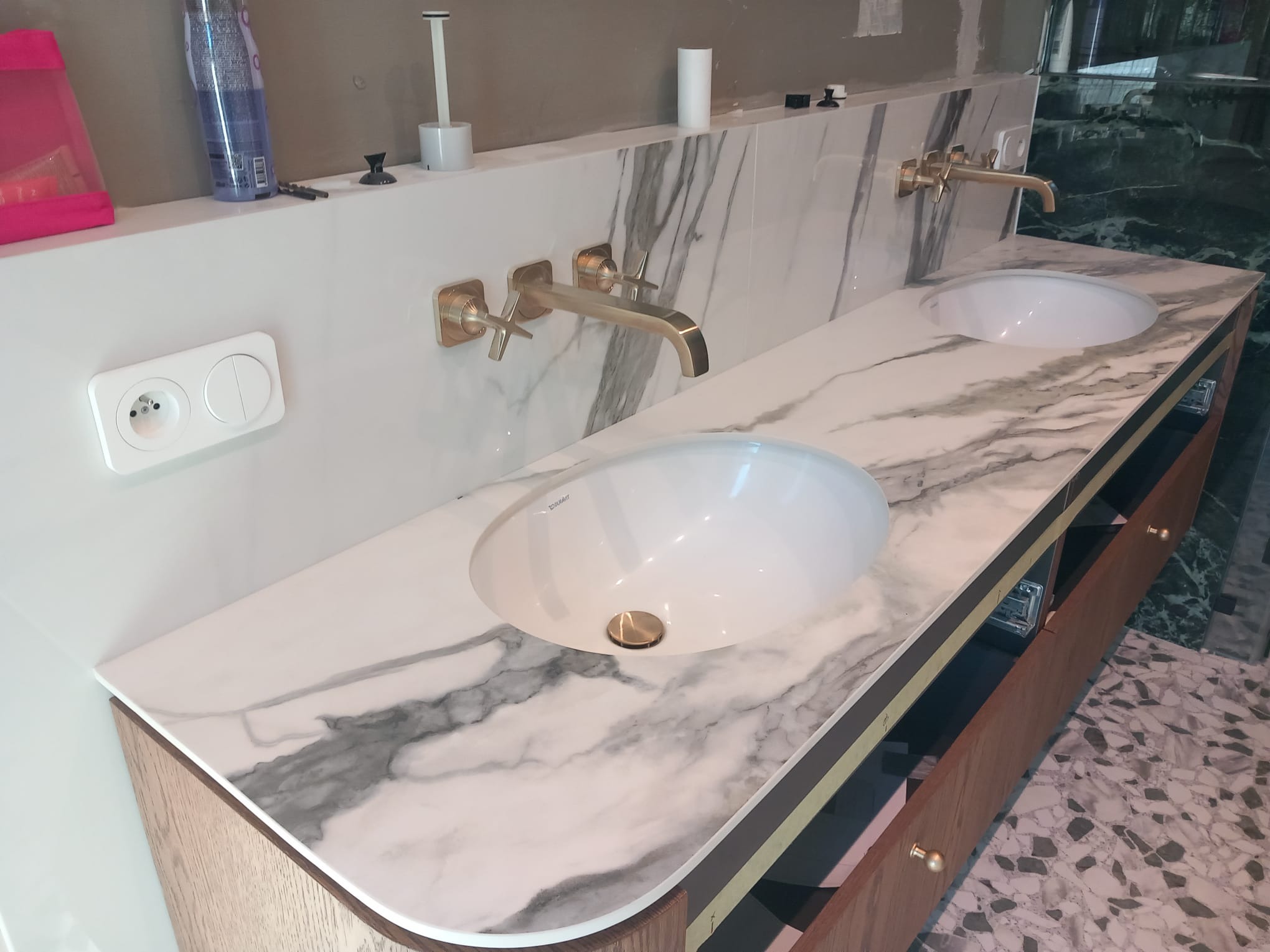 Marmurowy blat łazienkowy z dwoma umywalkami Duravit i złotą armaturą ścienną. Widoczne gniazdko elektryczne, półka i fragment podłogi z mozaiką.
