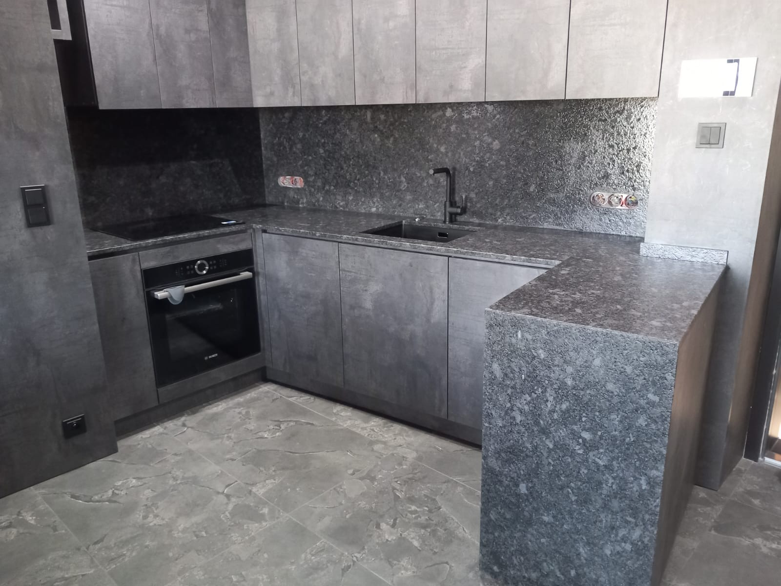 Nowoczesna kuchnia w odcieniach szarości: kamienny blat, zabudowa meblowa z piekarnikiem i płytą, ciemna armatura i płytki imitujące kamień na ścianie.