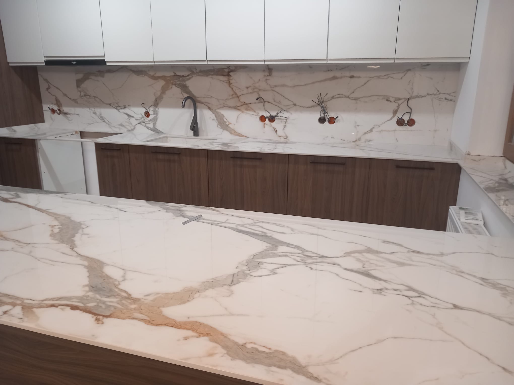 Biały blat kamienny z marmurowym wzorem w kuchni, brązowe szafki dolne, białe szafki górne, czarna bateria, widoczne instalacje elektryczne nad blatem.